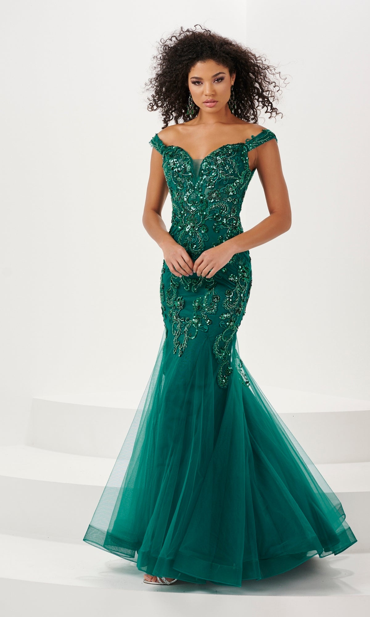 A woman stands against a white background wearing the Long Prom Dress 14185 by Panoply, an off-the-shoulder fitted emerald green mermaid gown with sequin embroidery and a tulle skirt.